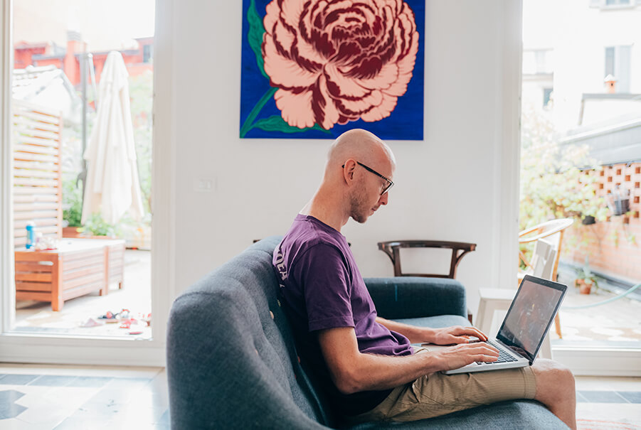 Homem adulto trabalhando home office no interior da casa sentado no sofá usando o computador.