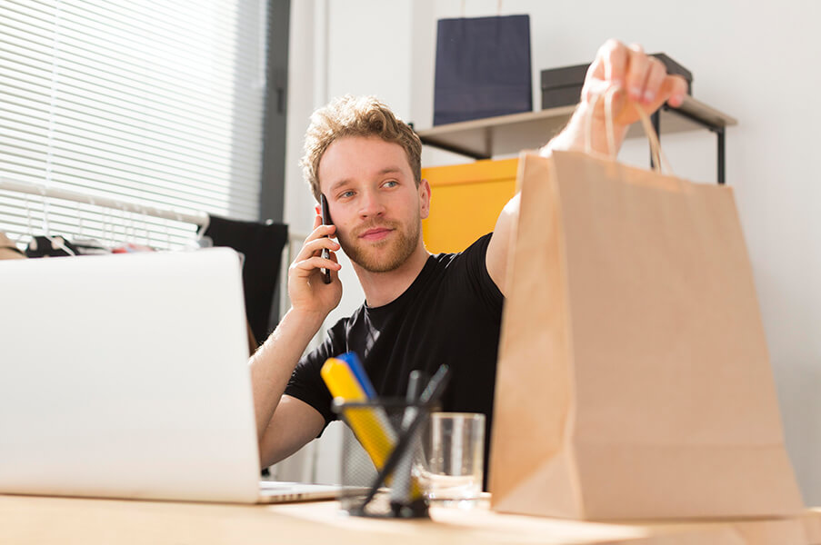 Homem em frente ao computador falando no celular e segurando uma sacola de papelão